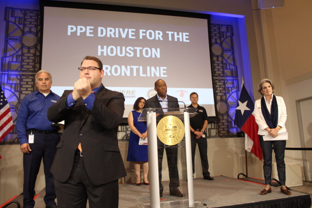 Mayor Sylvester Turner speaks at the podium in the City Hall Legacy Room during a news conference where he announced the "PPE drive for the Houston Frontline" (which is written on a screen behind him). Representatives from Project CURE, Cheniere Energy, and the Astros Foundation stand socially distant from him on stage. An American Sign Language Interpreter stands in the foreground. 